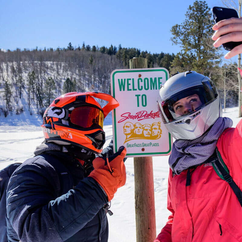 Two Black Hills Snowmobilers posing for a selfie in front of a welcome to South Dakota sign